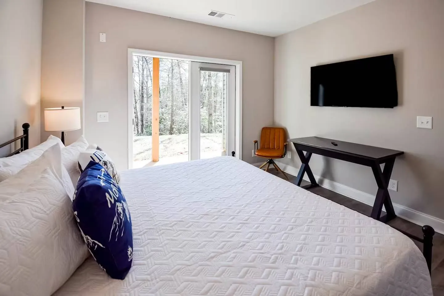 Bedroom with a white quilted bed, a wall-mounted TV, a small desk, and sliding glass doors leading outside.