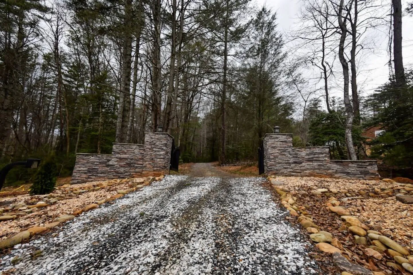 A gravel driveway flanked by stone pillars and river rocks leads into a wooded area.