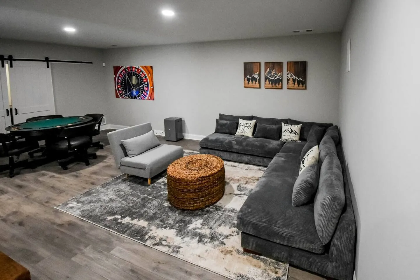 Game room and lounge featuring a large gray sectional sofa, woven ottoman, area rug, and poker table with black rolling chairs.