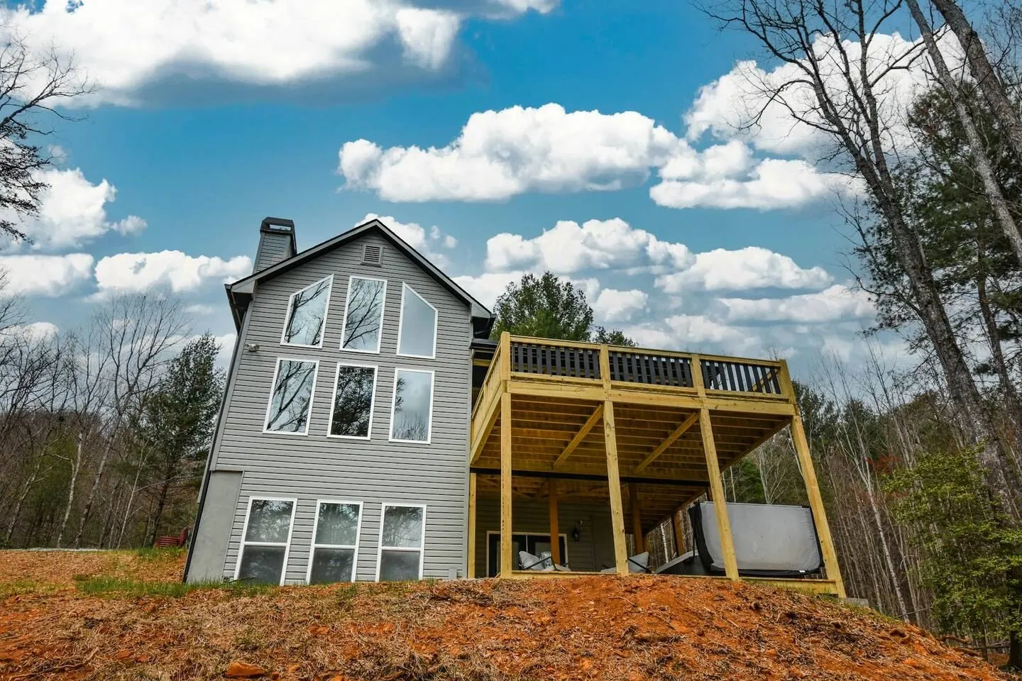 Rear exterior view of a multi-story gray siding house with large windows, an elevated wooden deck, and a hot tub on the covered patio below.