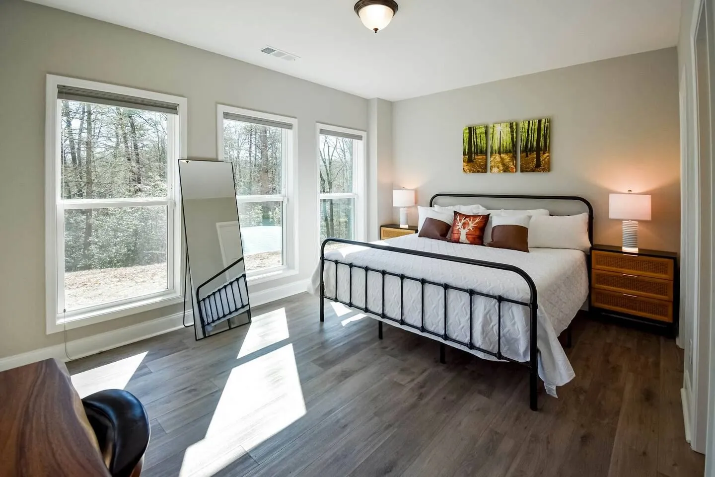 Bedroom with a black metal bed frame, nightstands with lamps, a full-length mirror, and large windows looking out onto trees.