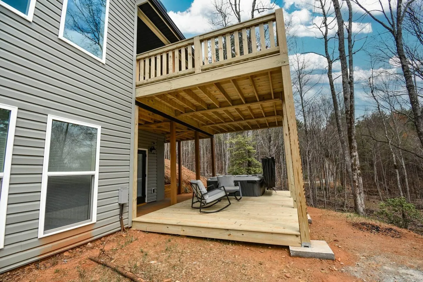 Rear exterior of a house featuring a two-story wooden deck with a lower patio holding rocking chairs and a hot tub.