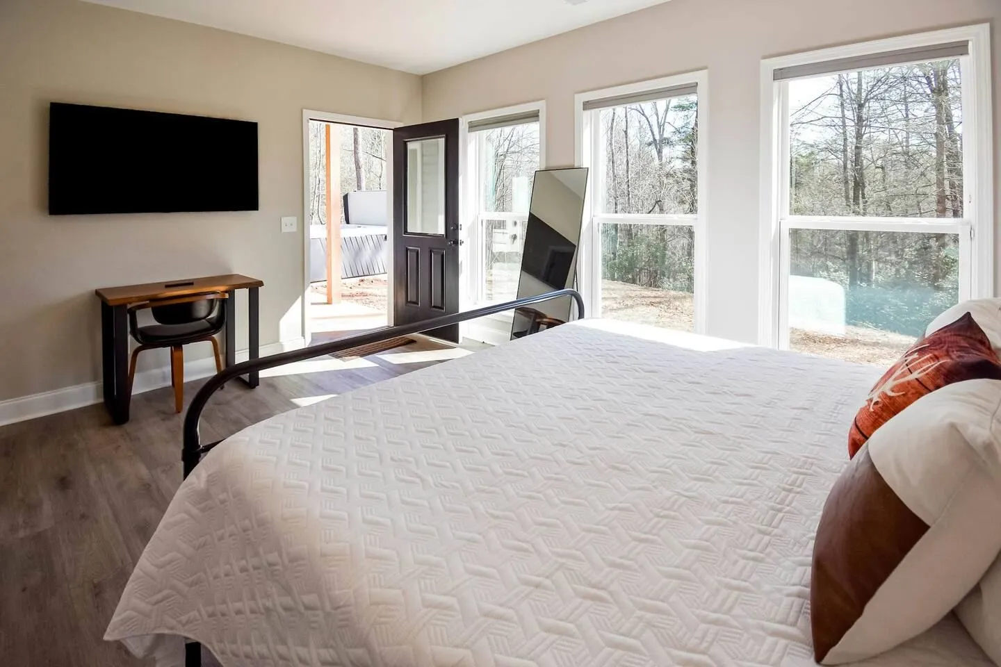 Bedroom with a white-covered bed, large windows, a wall-mounted TV, small desk, and an exterior door leading outside.