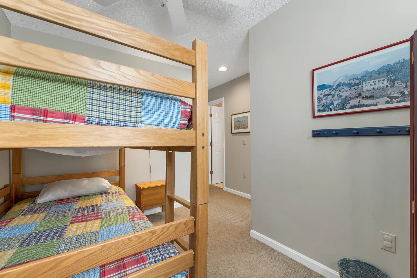 Bedroom featuring a wooden bunk bed with colorful patchwork quilts on carpeted flooring, looking toward a hallway doorway.