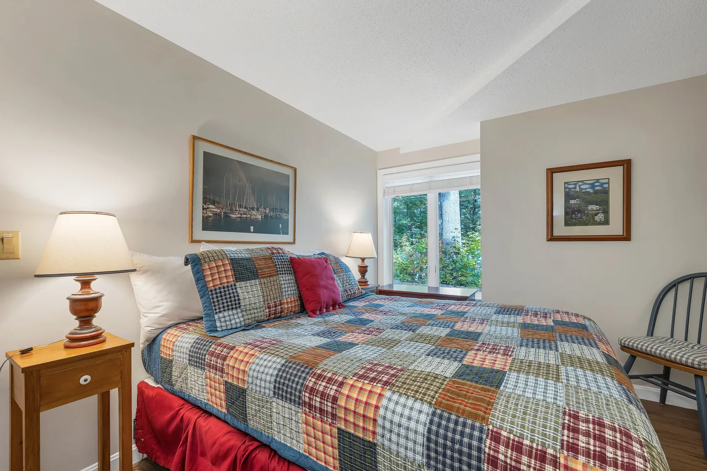 Bedroom with a patchwork quilt on the bed, bedside table with lamp, framed artwork, and a window showing outdoor greenery.