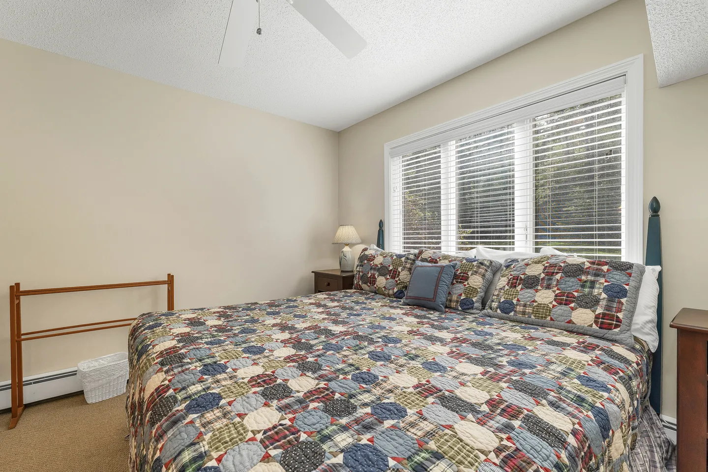 Bedroom with a patterned quilt-covered bed, nightstands, and a large window with white horizontal blinds.