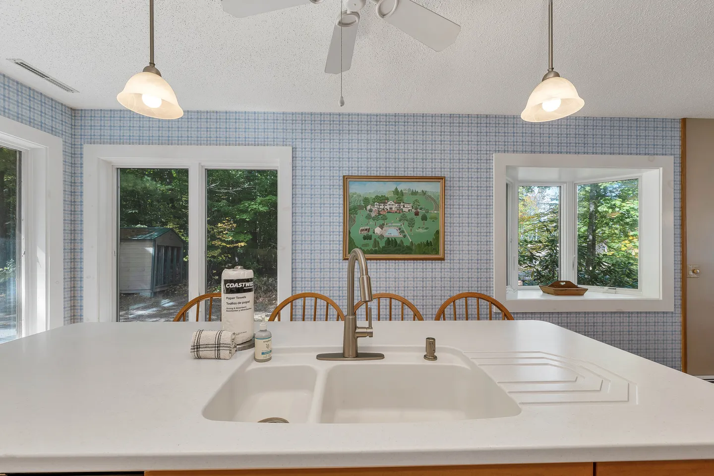 Kitchen island with a white double sink and faucet in the foreground, facing wooden dining chairs, blue patterned wallpaper, and windows overlooking greenery.