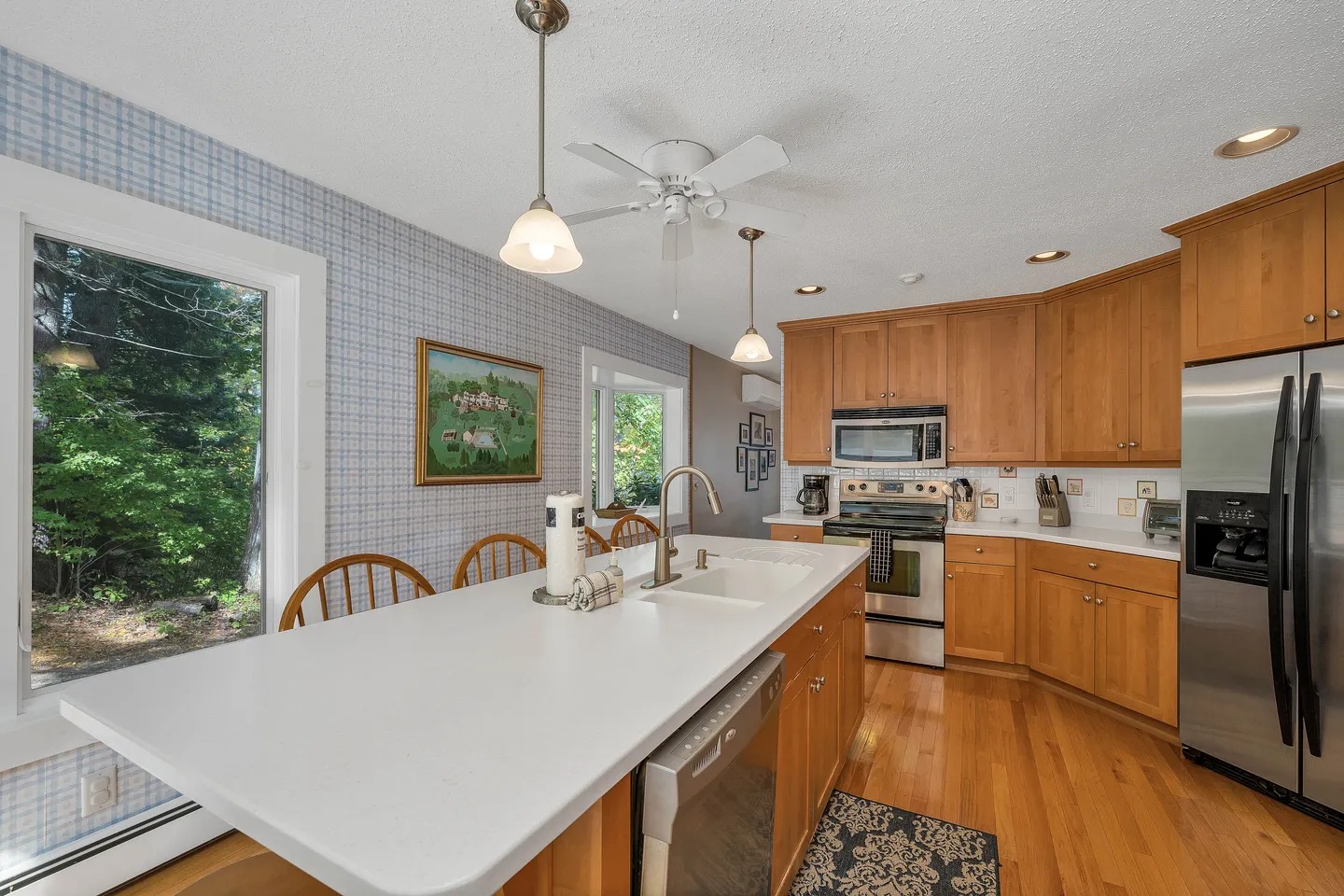 Kitchen featuring a large white center island with a built-in sink and dishwasher, wood cabinetry, stainless steel appliances, and a window viewing lush greenery.