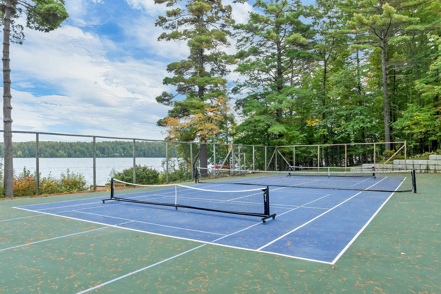 Outdoor sports court with pickleball nets set up beside a lake surrounded by tall pine trees.