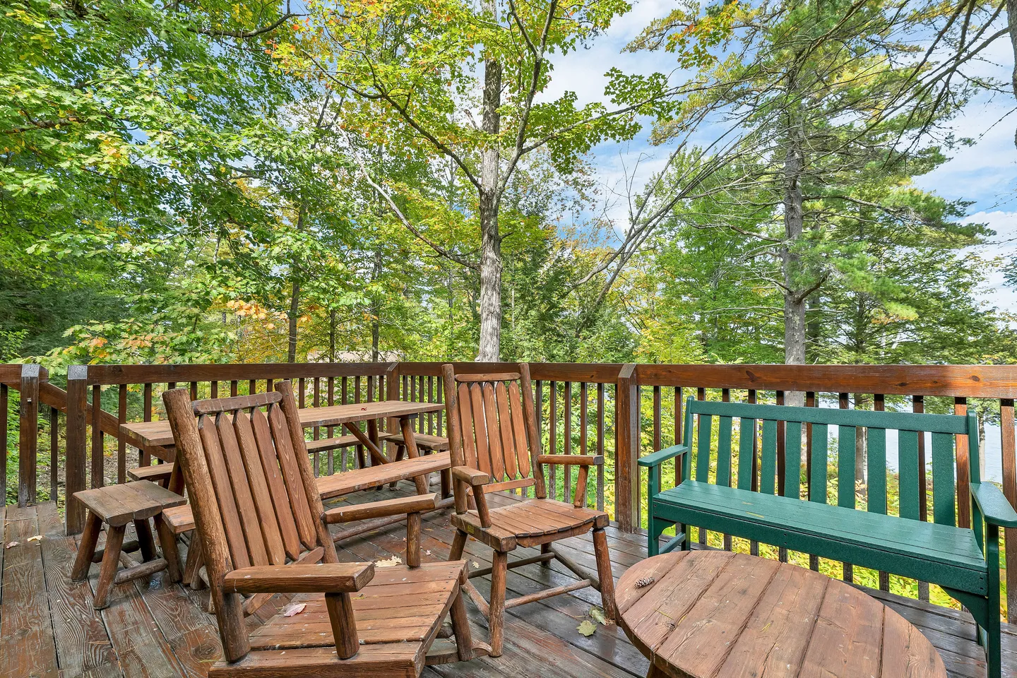 Outdoor wooden deck with rustic wooden armchairs, a green bench, and a picnic table surrounded by lush green trees.