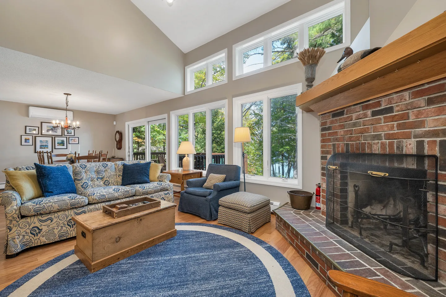 Living room with vaulted ceiling, large windows overlooking trees, a brick fireplace, patterned sofa, and blue armchair.