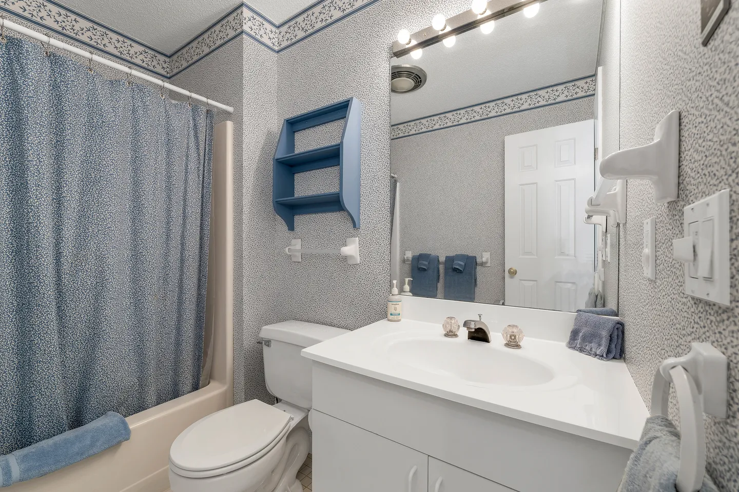 Bathroom featuring a white vanity with mirror, a toilet, patterned wallpaper with border, and a bathtub with a blue shower curtain.