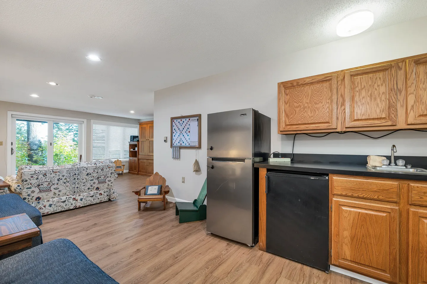 Open living area with wood-look flooring, featuring a kitchenette with wooden cabinets and a refrigerator next to a living space with sofas and a sliding glass door.