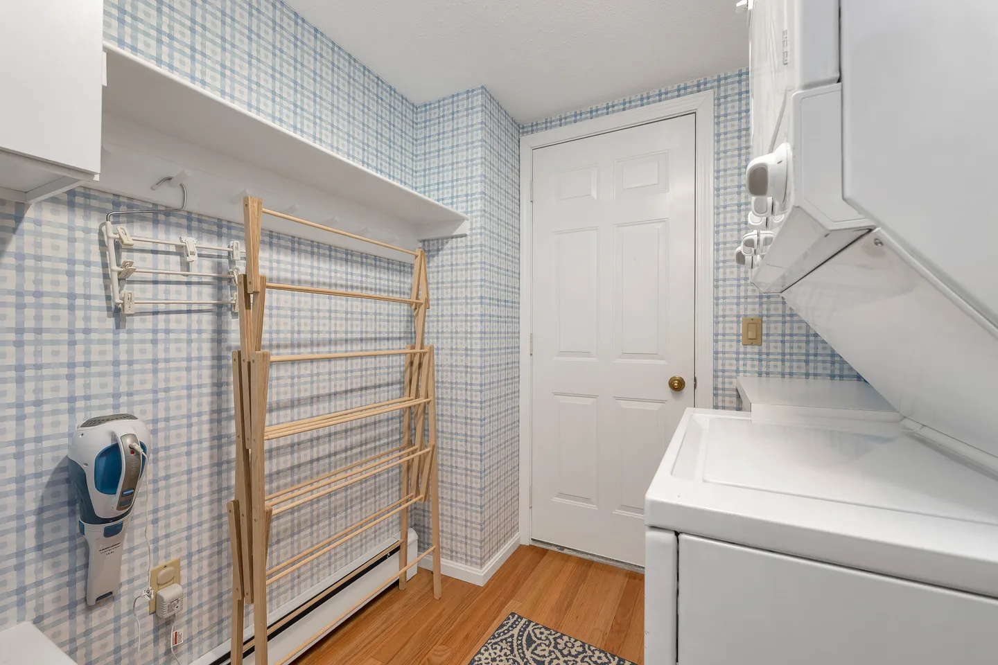 A laundry room featuring light blue plaid wallpaper, a stacked washer and dryer, and a wooden drying rack.