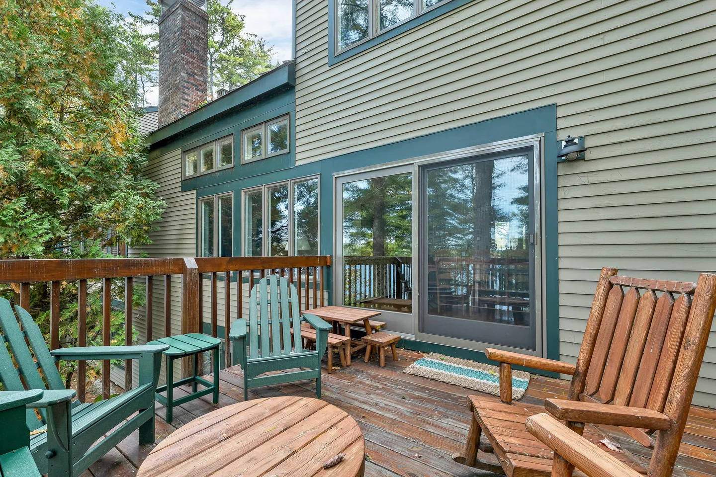 Wooden deck with Adirondack chairs, a small picnic table, and sliding glass doors leading into a green-sided house.