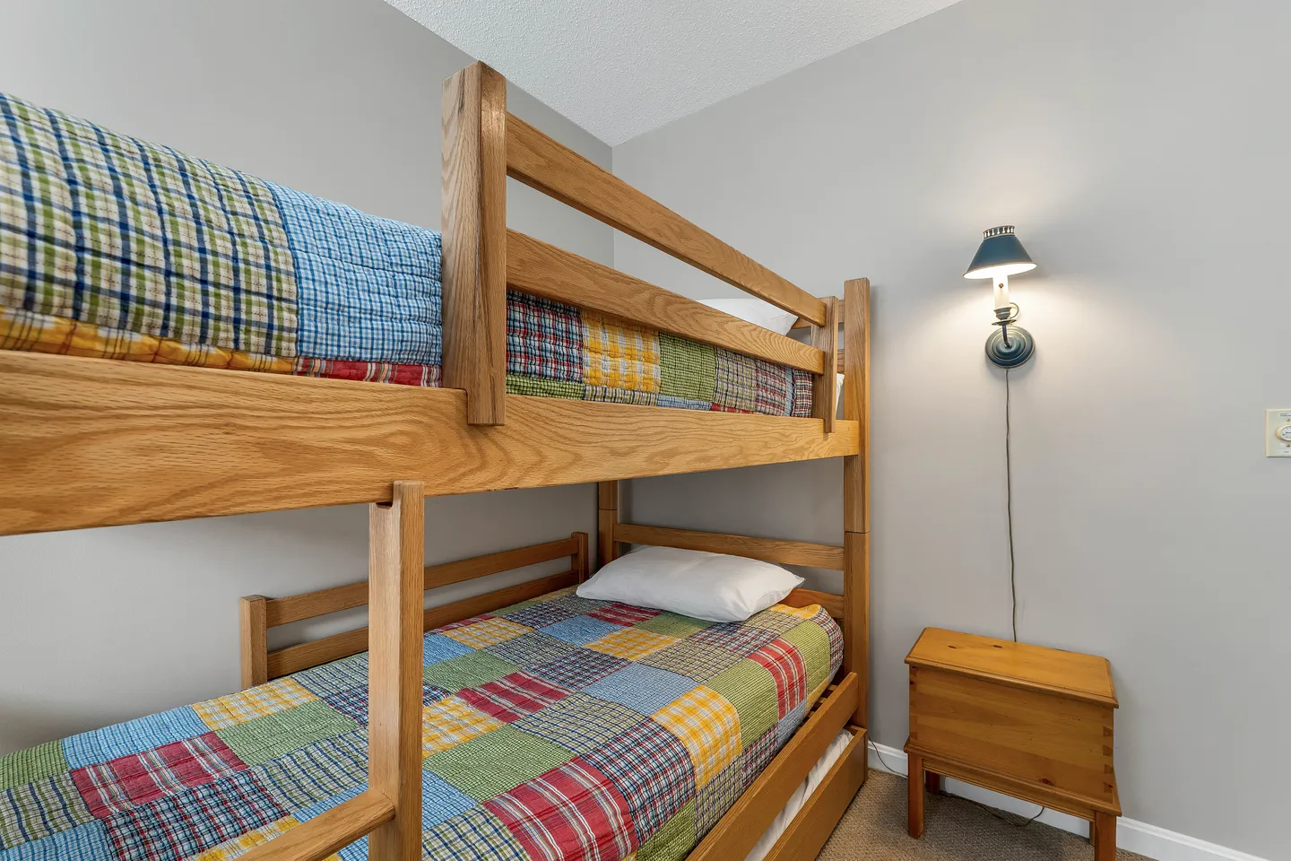 Wooden bunk bed with colorful patchwork quilts beside a small side table and wall-mounted lamp.