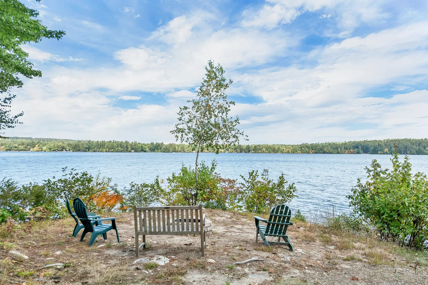 Outdoor seating area with a wooden bench and plastic chairs overlooking a wide lake under a partly cloudy sky.