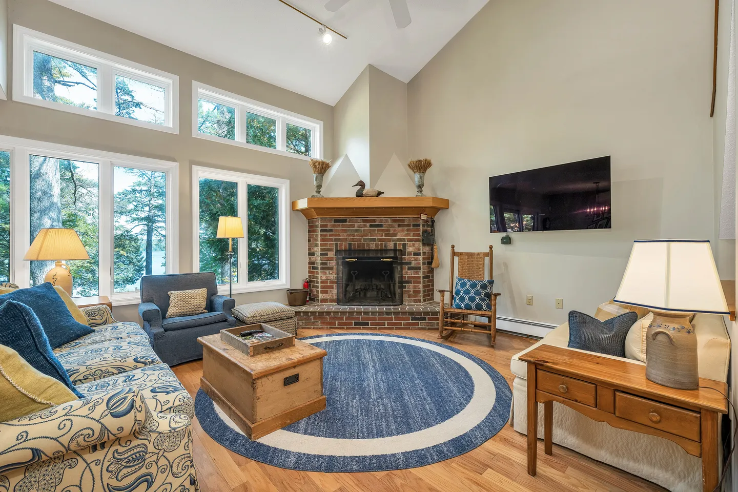 Living room featuring a corner brick fireplace, vaulted ceilings, large windows overlooking trees, patterned couches, and a round blue rug.