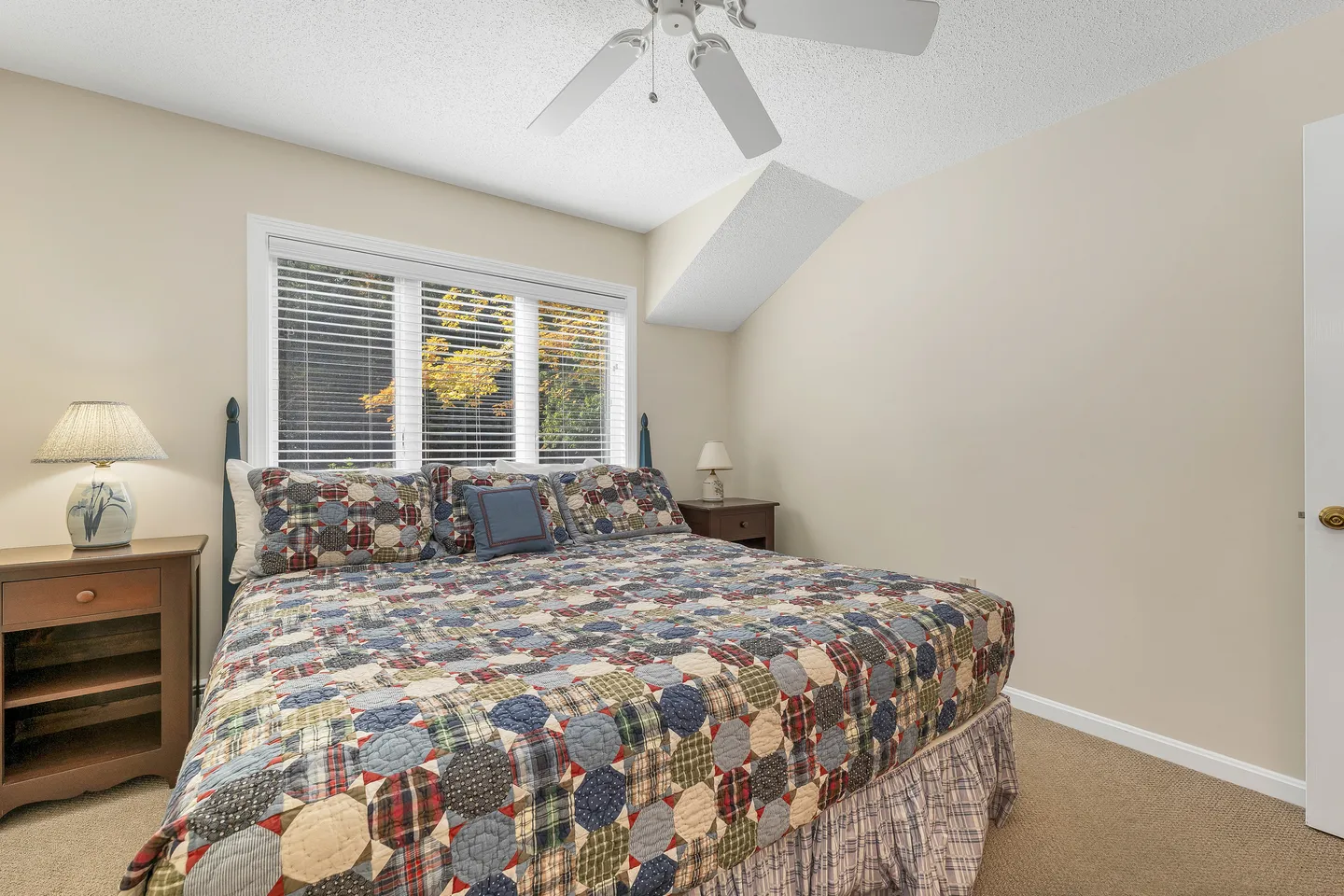 Bedroom with a patterned quilt on a bed flanked by nightstands and a window with horizontal blinds.