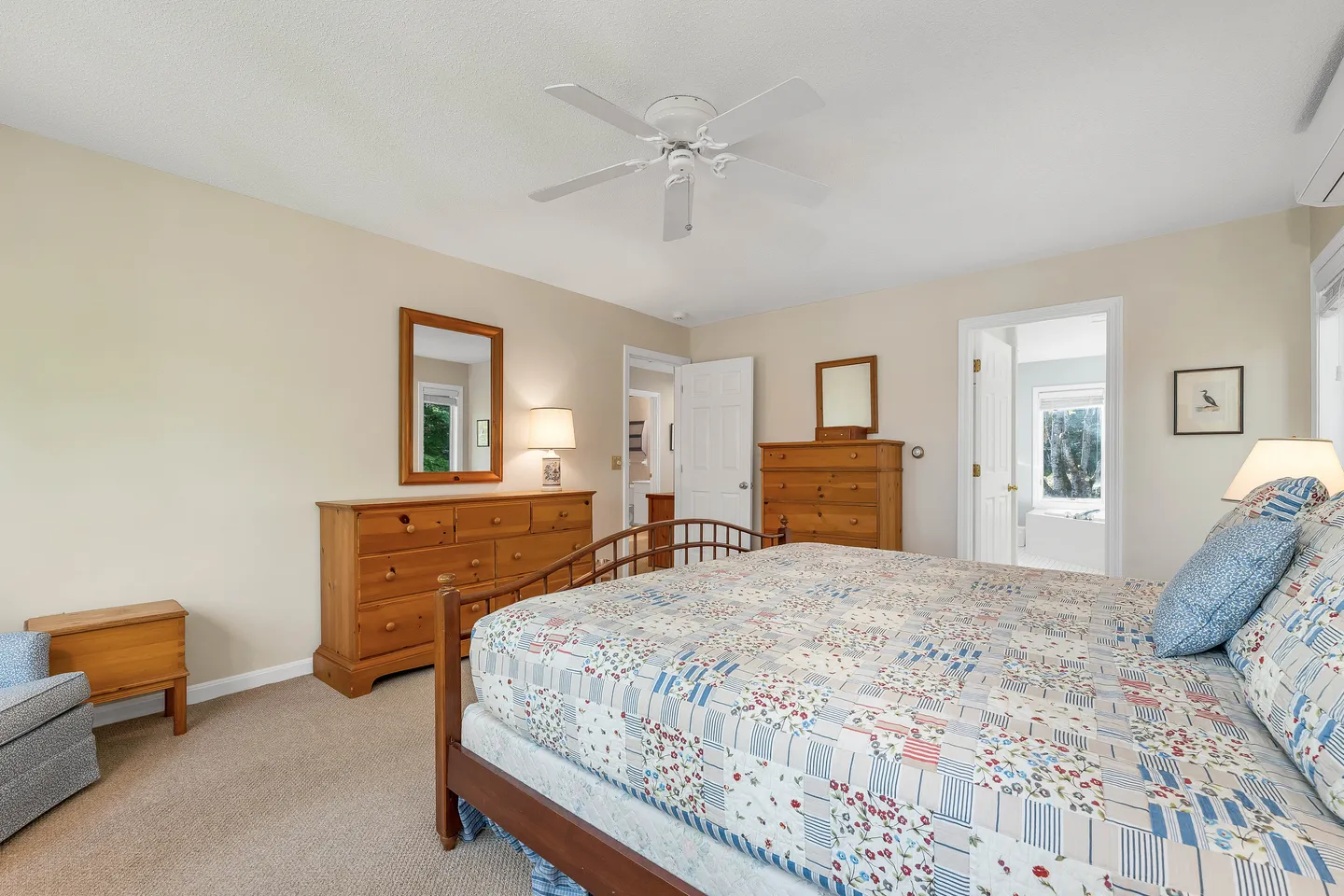 Bedroom with a wooden bed, patterned quilt, dressers, ceiling fan, carpet, and an open doorway leading to a bathroom.