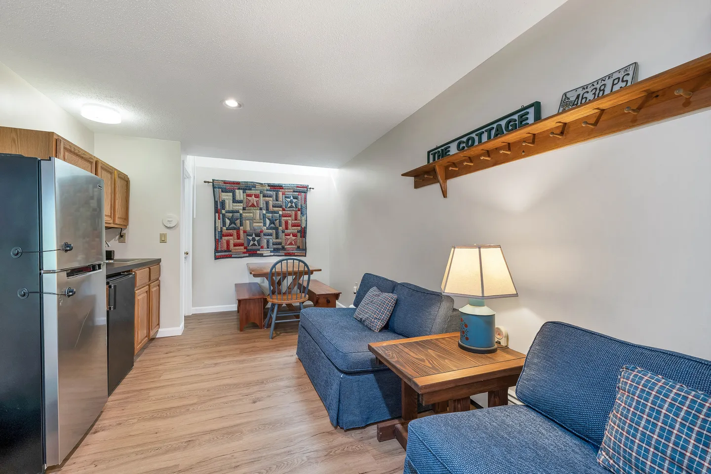 Open living area with blue upholstered seating, wood-look flooring, kitchen cabinets, and a dining table with a quilt wall hanging.