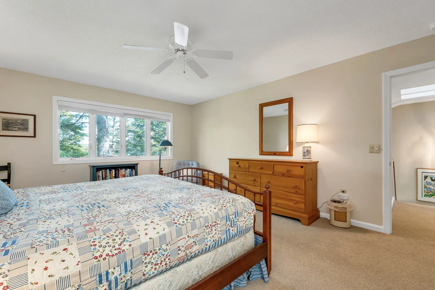 Bedroom with carpet flooring, a wooden bed topped with a patchwork quilt, wooden dresser, ceiling fan, and large window overlooking trees.