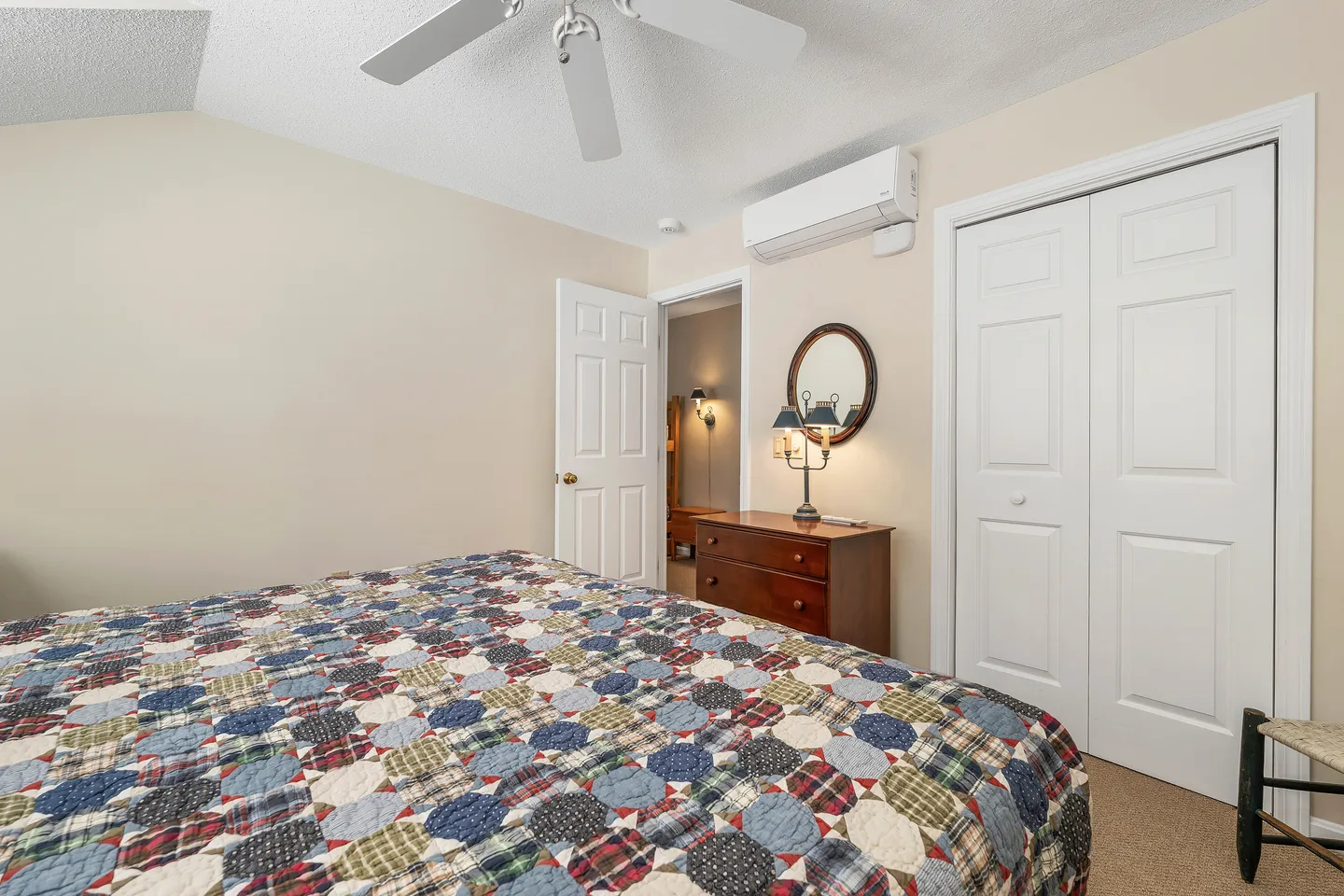 Bedroom with a colorful patchwork quilt on the bed, a wooden dresser with a mirror, a ceiling fan, and a mini-split AC unit.