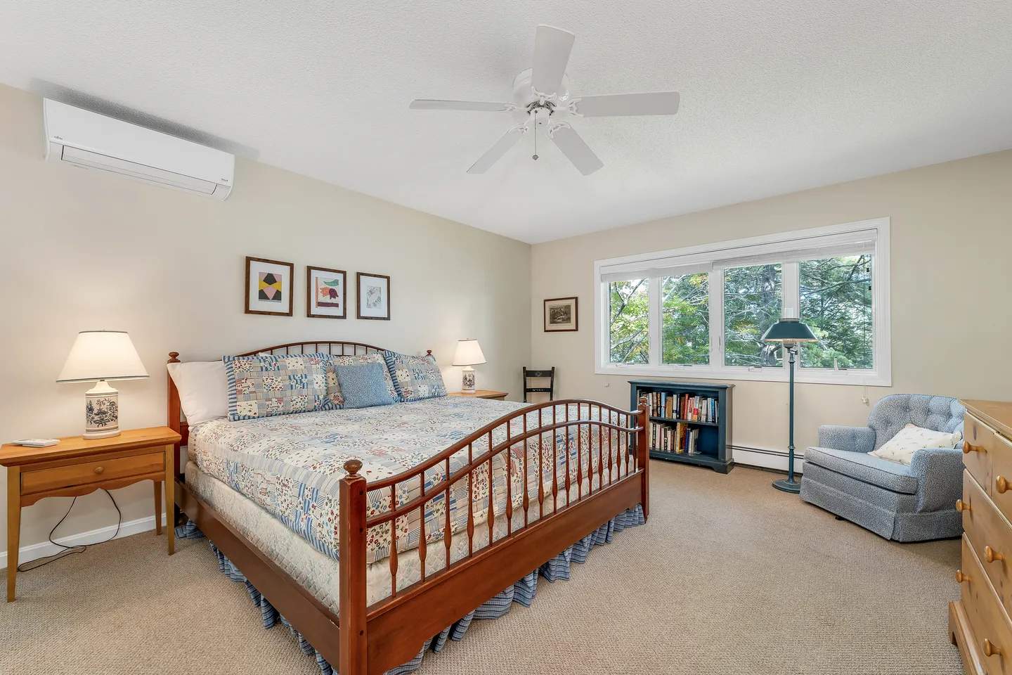 Bedroom with wooden spindle bed, beige carpet, ceiling fan, wide window overlooking trees, and an armchair next to a low bookcase.