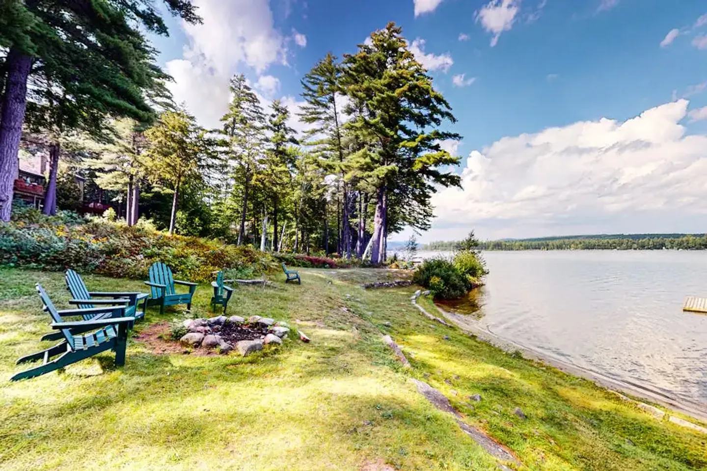 Lakeside lawn with green Adirondack chairs arranged around a stone fire pit near mature pine trees and the water's edge.