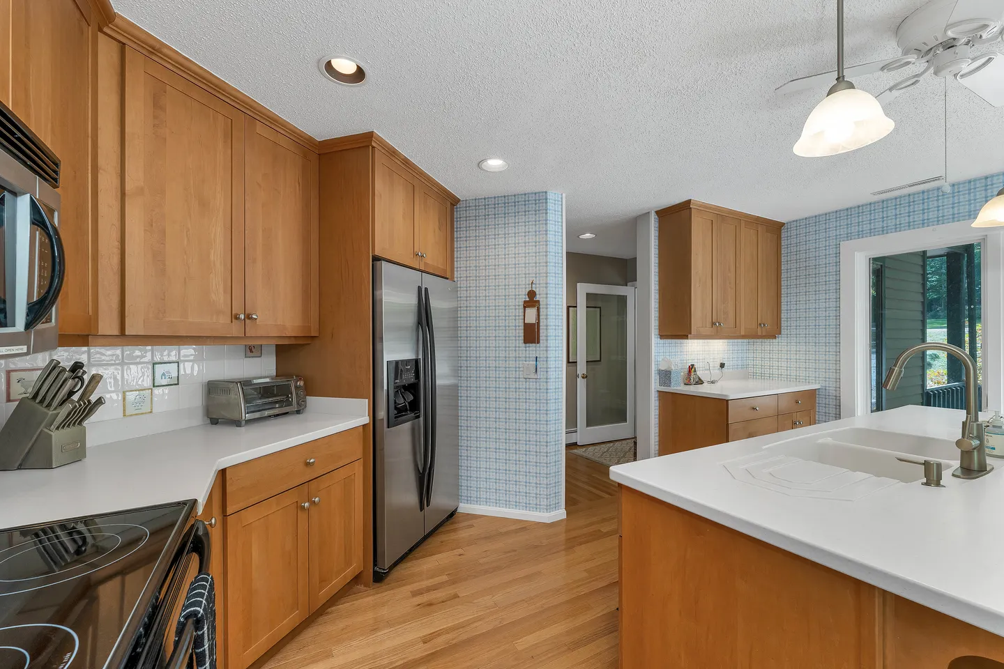Kitchen featuring light wood cabinets, white countertops, stainless steel appliances, light wood flooring, and light blue patterned wallpaper.