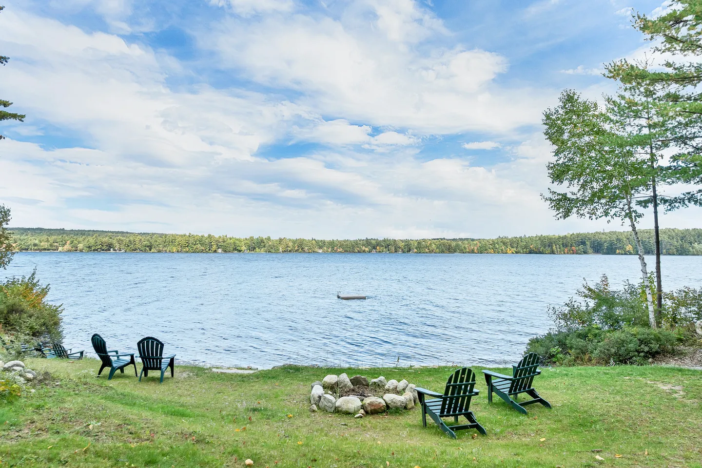 Outdoor lawn by a lake featuring dark green Adirondack chairs and a stone fire pit overlooking the water.