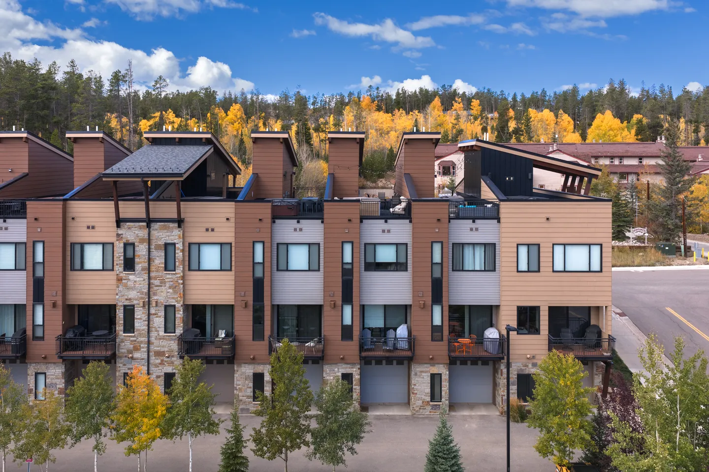 Exterior view of modern multi-level townhomes with balconies, stone accents, and autumn trees against a forested hill.