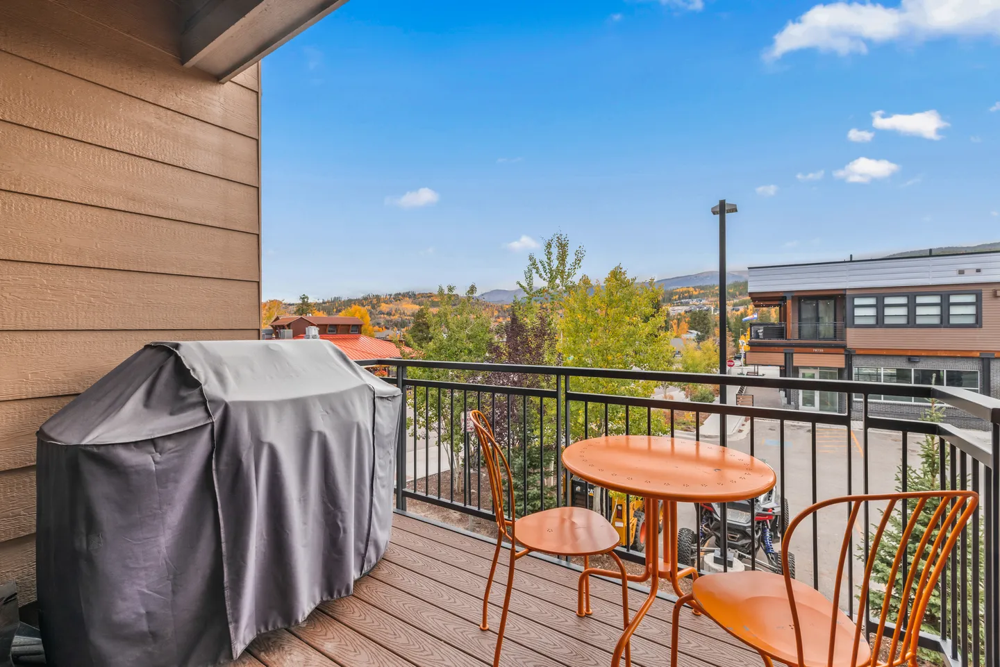 Balcony with covered grill and orange metal patio table and chairs overlooking surrounding buildings and trees under a blue sky.