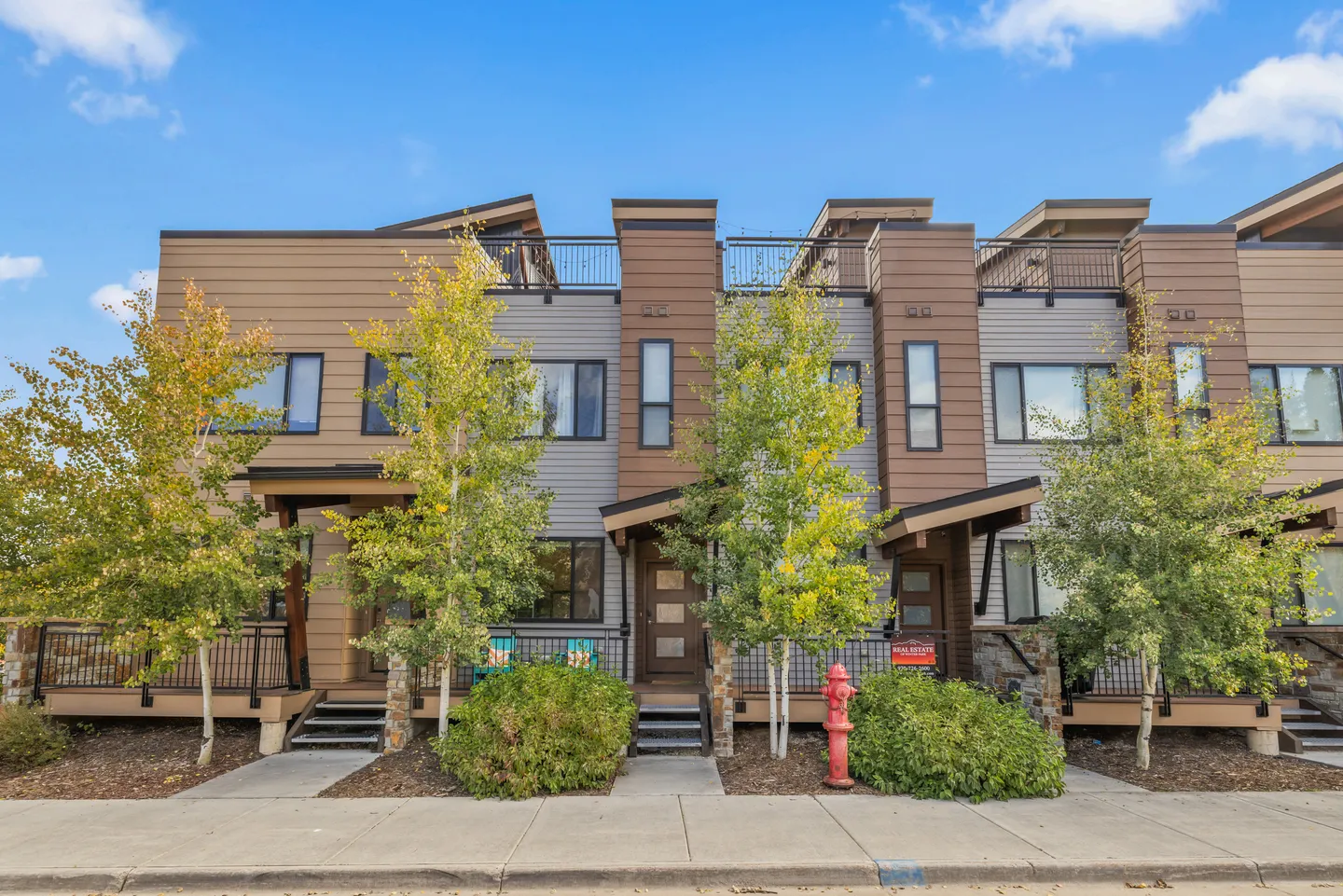 Exterior front facade of modern multi-level townhomes with wood and gray siding, front steps, small trees, and a sidewalk under a blue sky.