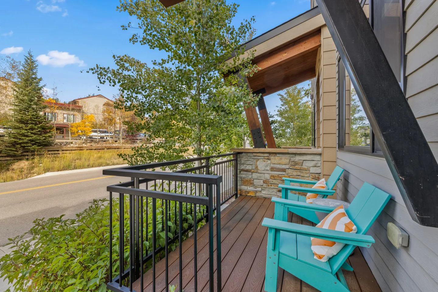 Covered outdoor porch with two turquoise Adirondack chairs and black metal railing overlooking a paved street.