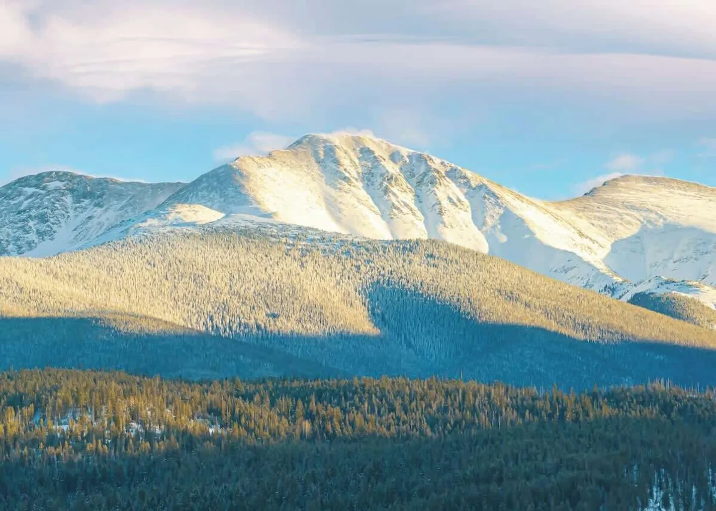 Snow-capped mountain peaks rising behind forested hills under a soft blue sky.