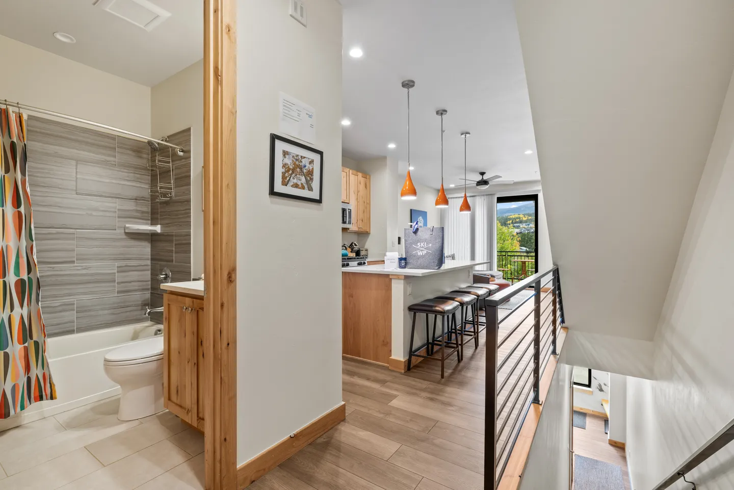 Hallway view showing a bathroom with a tub and vanity on the left, leading to an open-plan kitchen and living space, with a staircase visible on the right.