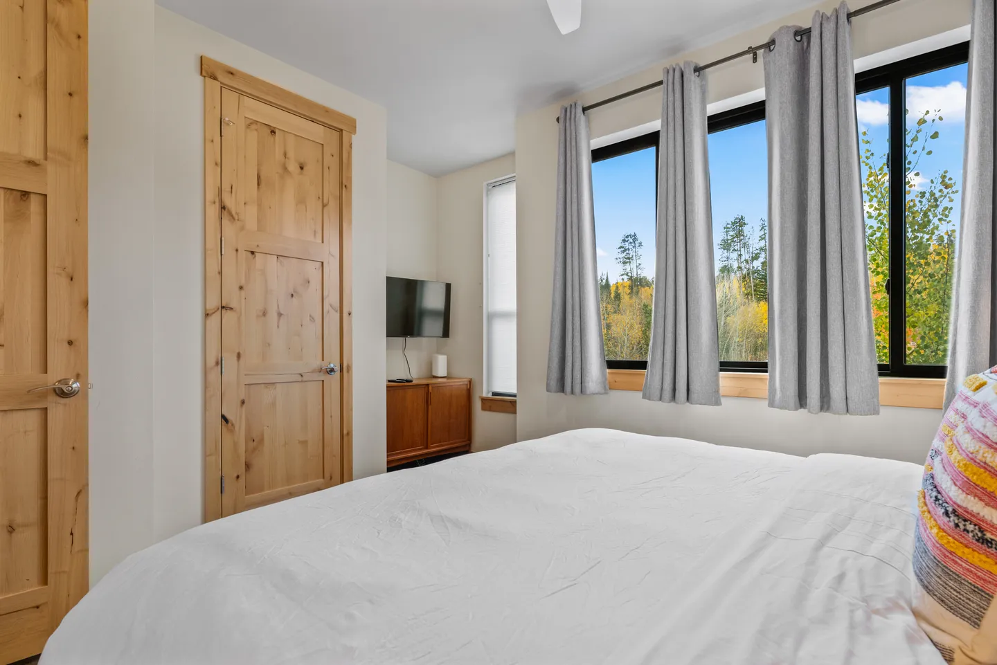 Bedroom with a white-linened bed, wood interior doors, and a wall of windows framed by grey curtains overlooking trees.