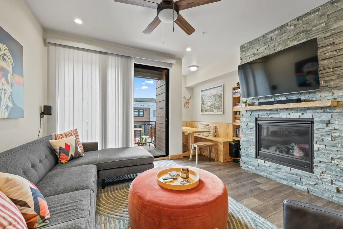 Living room featuring a grey sectional sofa, an orange round ottoman, a stone-faced fireplace with a wall-mounted TV, and sliding glass doors leading to a balcony.