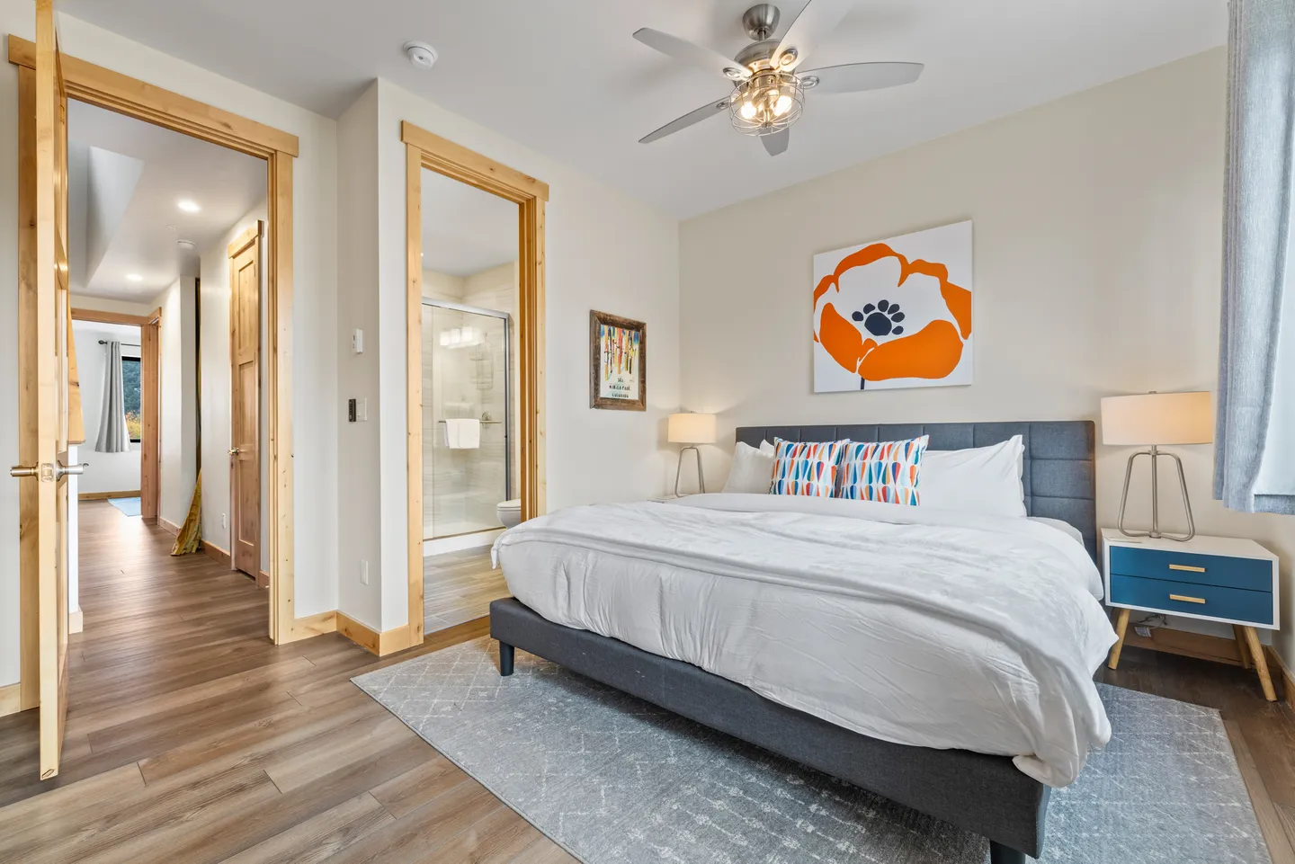 Bedroom with an upholstered bed, ceiling fan, bedside tables with lamps, and doorways leading to an en-suite bathroom and a hallway.