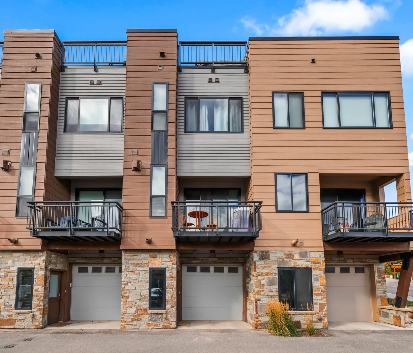 Exterior view of modern multi-story townhomes with stone accents, siding, garage doors, and private balconies under a blue sky.