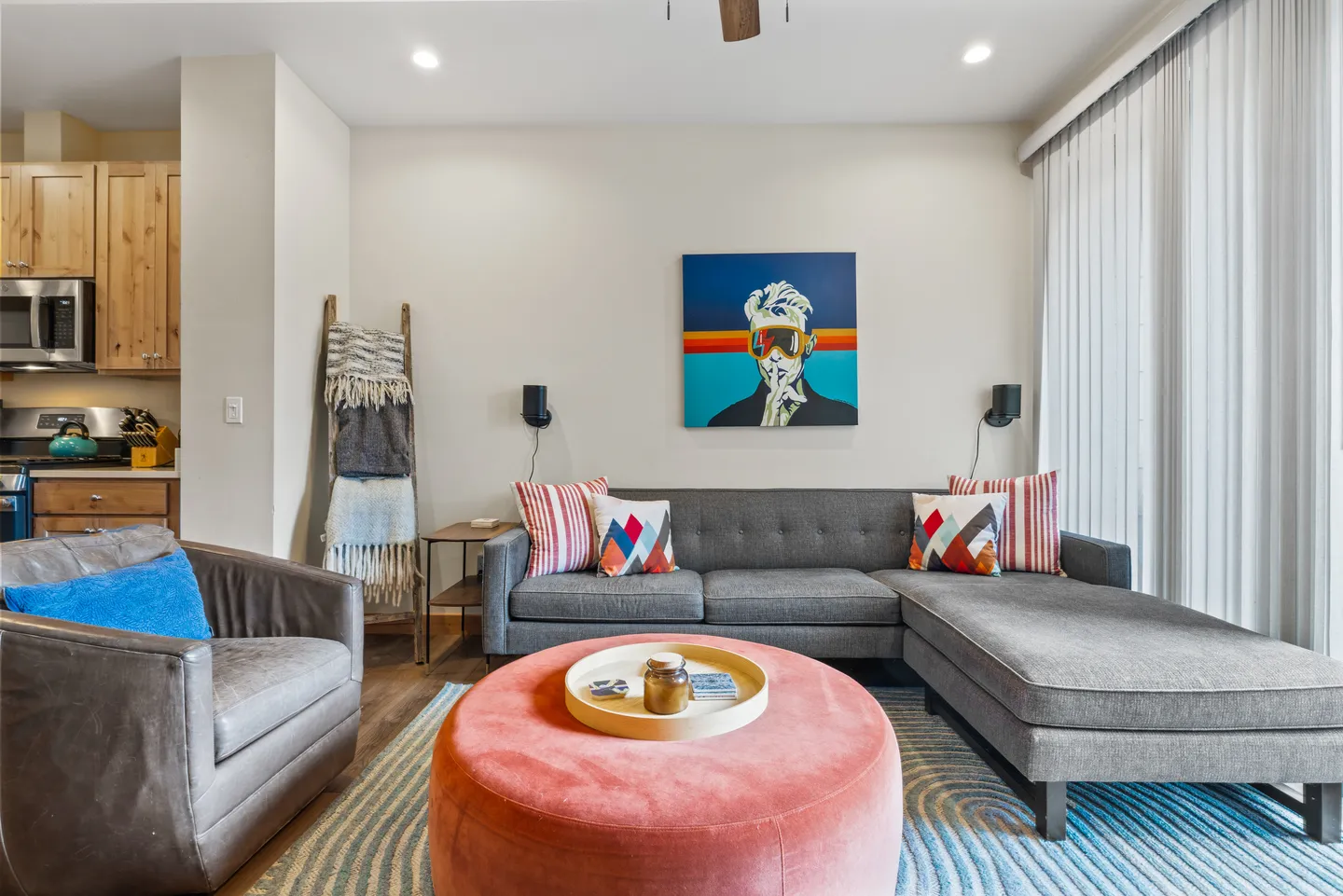 Living room featuring a grey sectional sofa with accent pillows, a large pink ottoman, an armchair, and artwork on the wall.