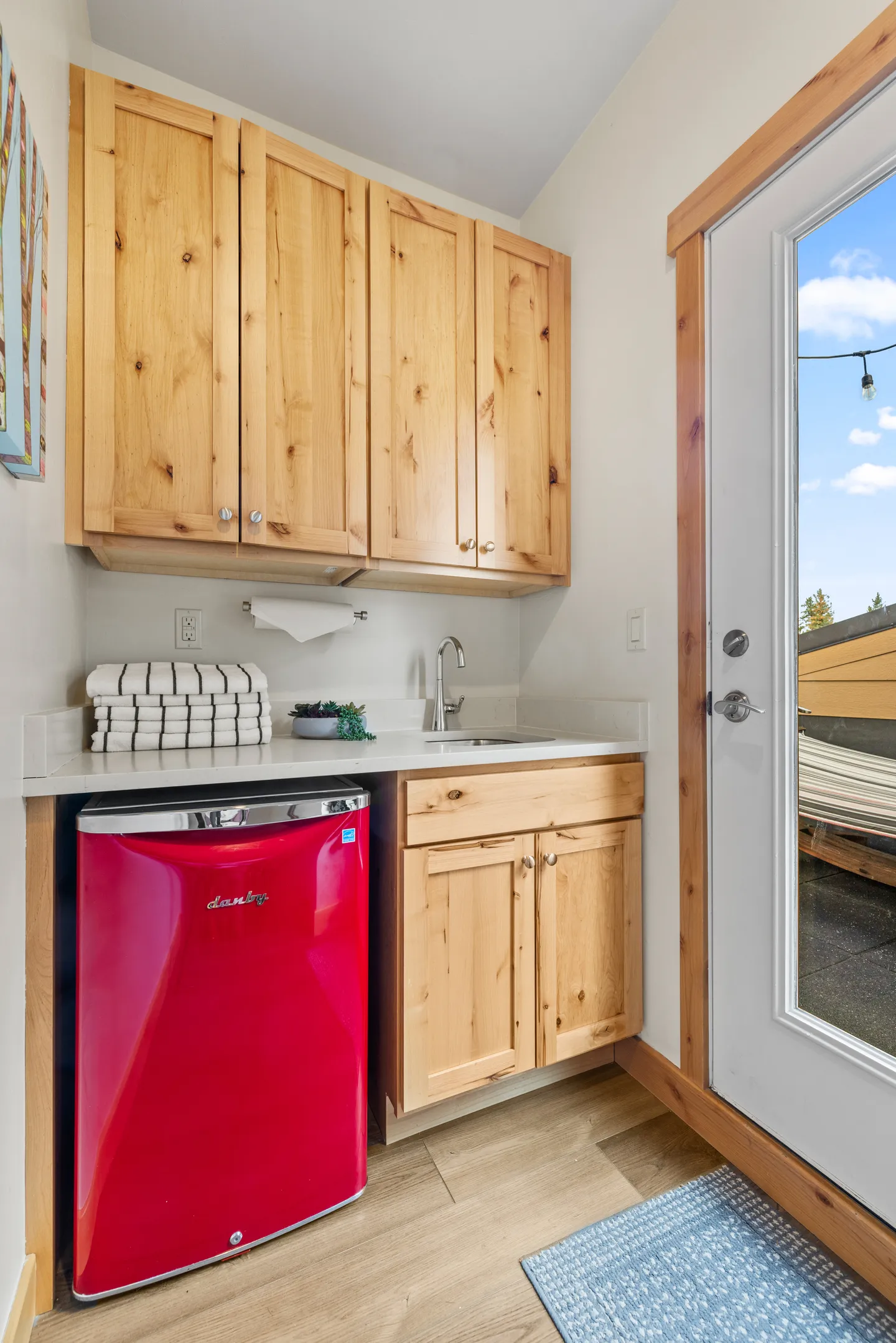 A kitchenette nook featuring light wood cabinets, a small sink, and a bright red mini fridge beside an exterior glass door.