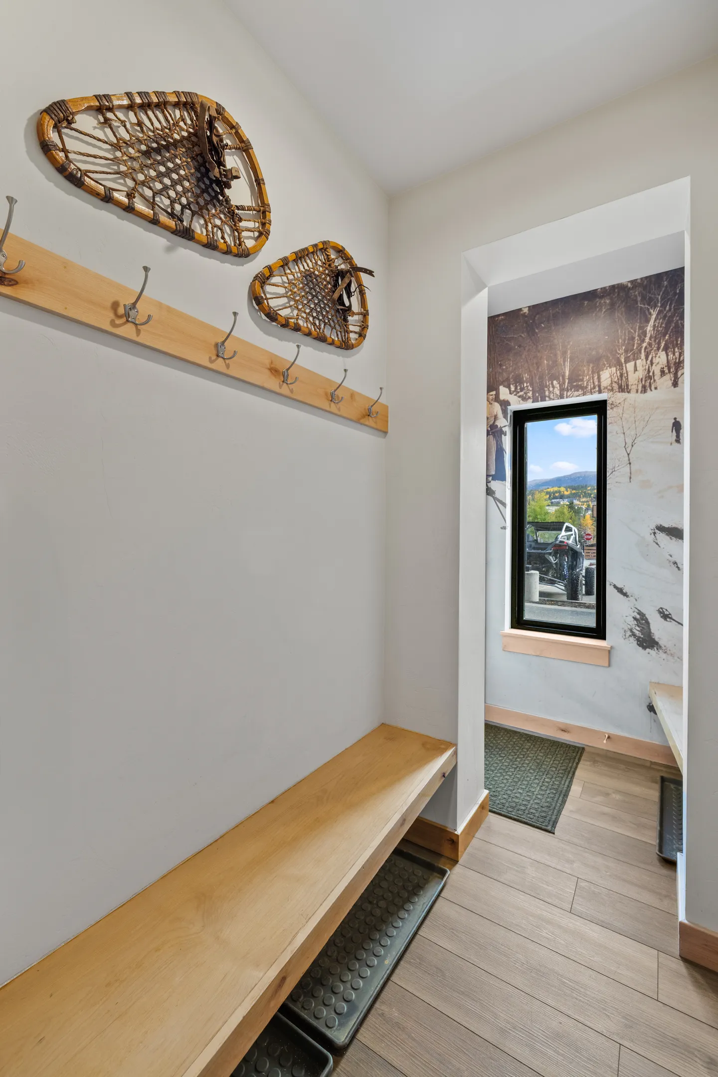 Mudroom entry area with a built-in wooden bench, coat hooks on a wooden board, decorative snowshoes, and a view into a windowed alcove with ski-themed wallpaper.