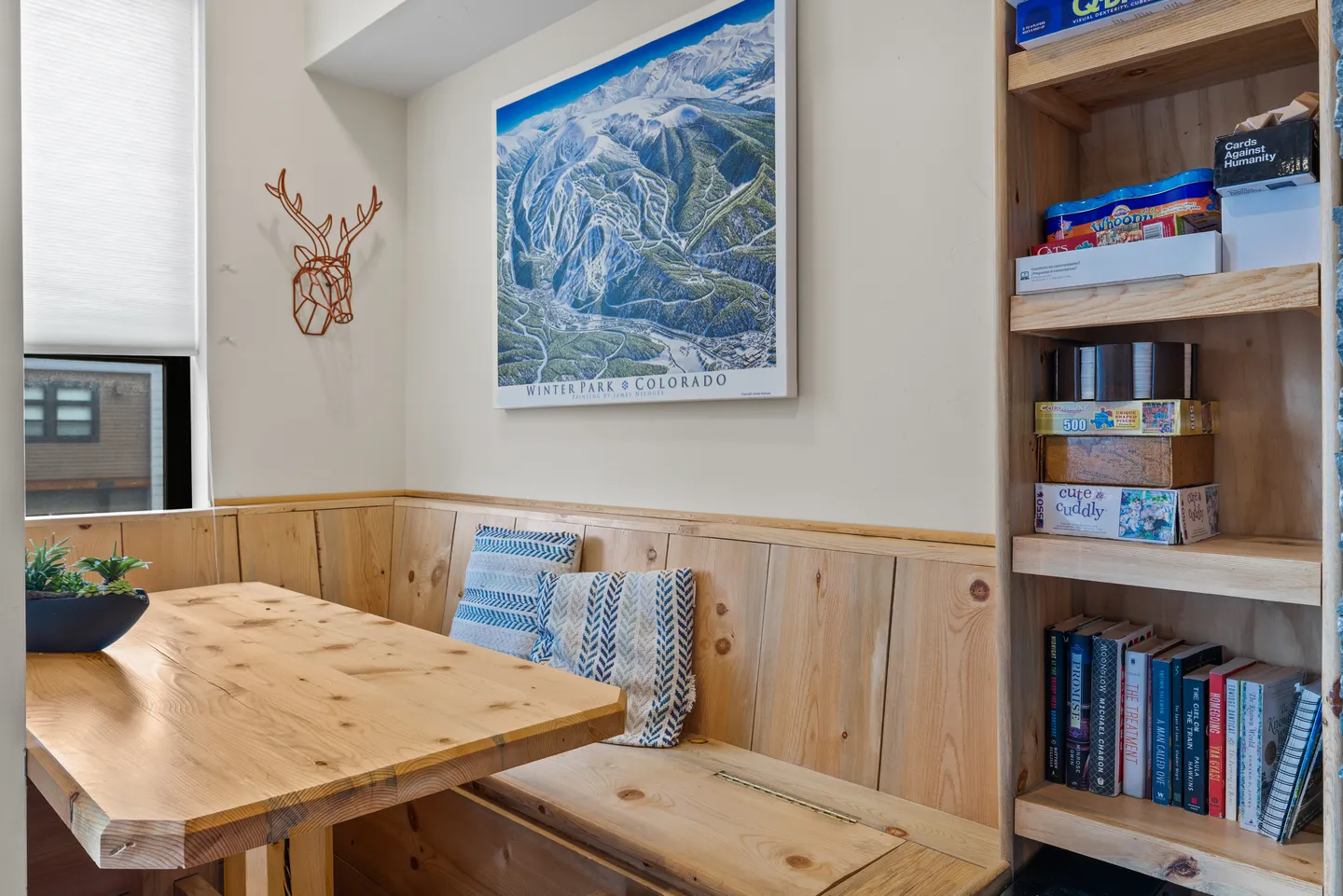 Built-in wooden dining nook with a table, bench seating, throw pillows, a bookshelf, and a framed mountain poster.