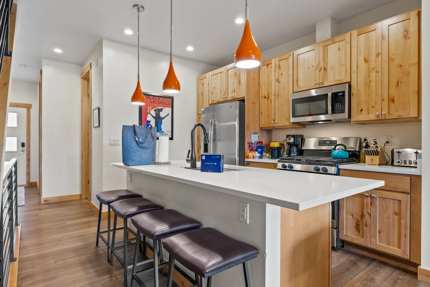 A kitchen featuring light wood cabinetry, stainless steel appliances, and an island with bar stools and orange pendant lights.