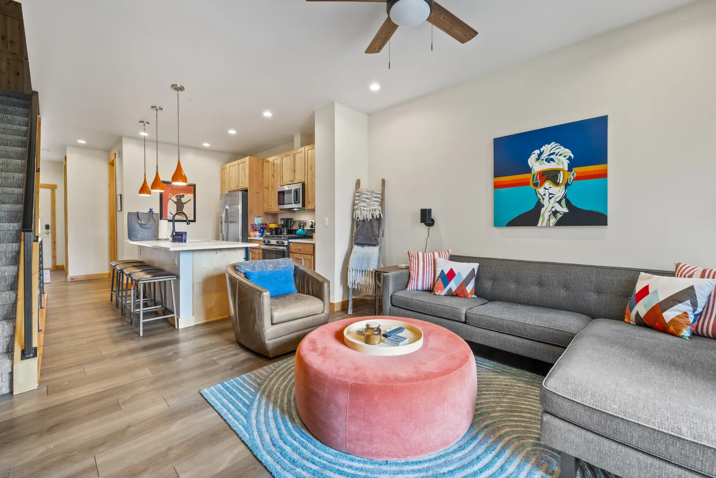 Open-concept living room with a sectional sofa and round pink ottoman, flowing into a kitchen with island seating.