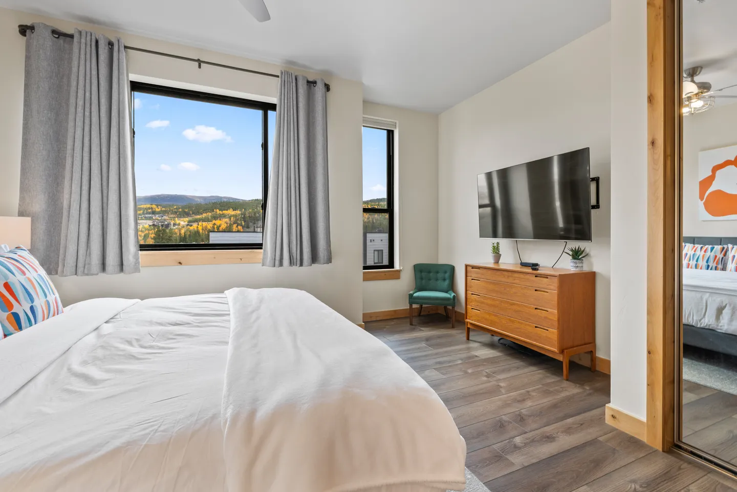 Bedroom with a white-dressed bed, windows showing mountain and forest views, a wall-mounted TV, and a dresser.