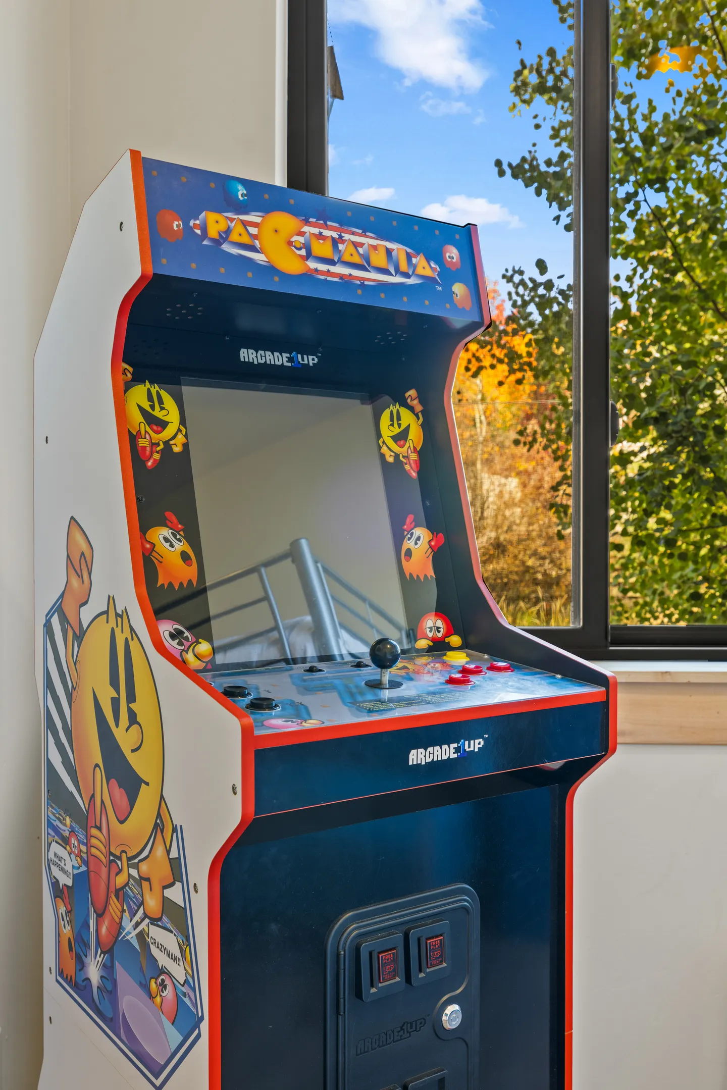 A Pac-Mania Arcade1Up gaming cabinet positioned next to a window showing outdoor trees and foliage.