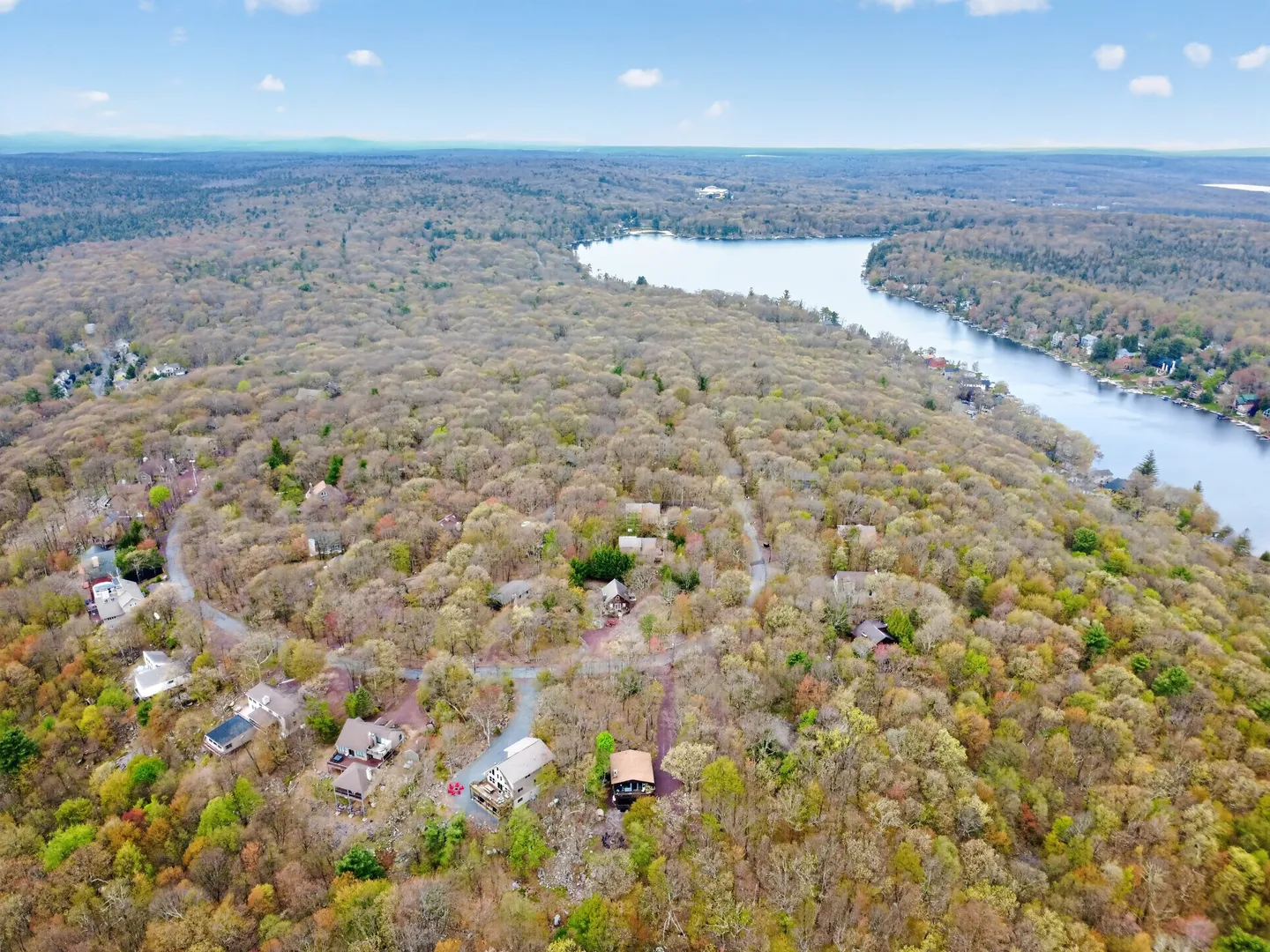 High-angle aerial photograph of a forested neighborhood with houses nestled among trees near a large lake under a blue sky.